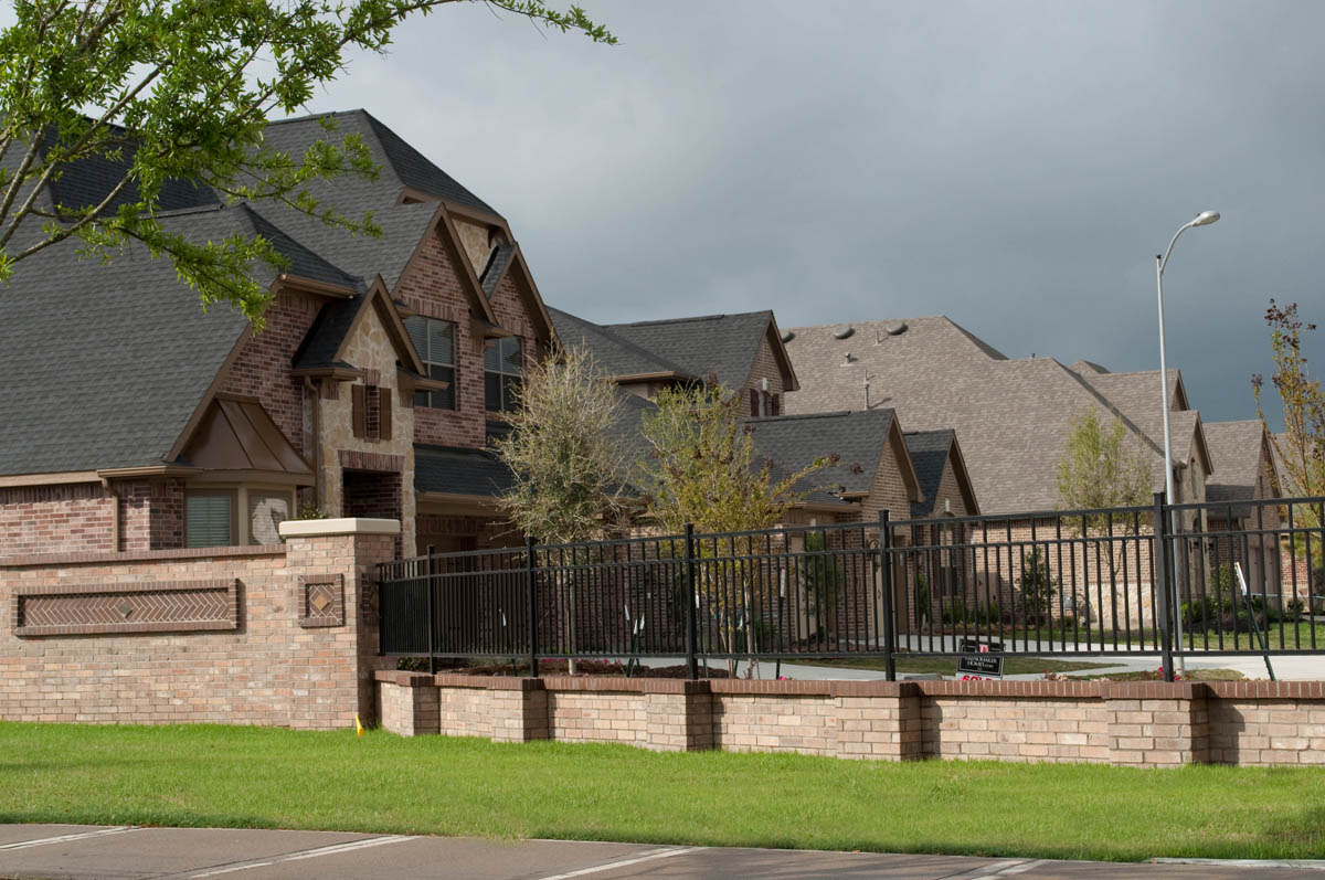 Row of modern suburban brick houses with dark roofs behind a black metal fence and brick wall, under a cloudy sky, with green grass and some trees in the foreground.