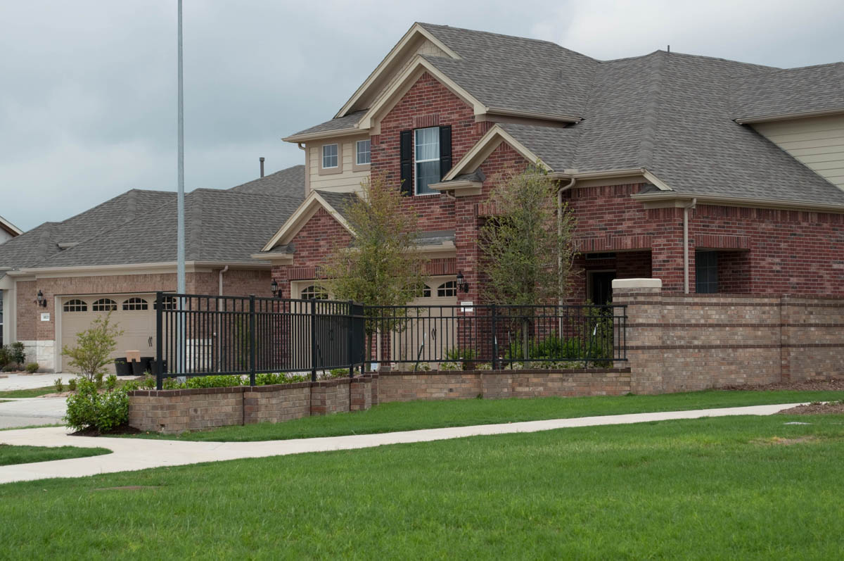 A two-story suburban brick house with a front yard, black metal fence, and a driveway leading to a two-car garage. The sky is cloudy and the lawn is green and well-kept.