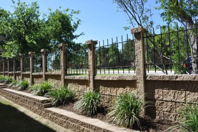 A tan stone retaining wall topped with vertical black metal railings runs alongside a planted bed with green ornamental grasses, with trees and a road visible in the background.