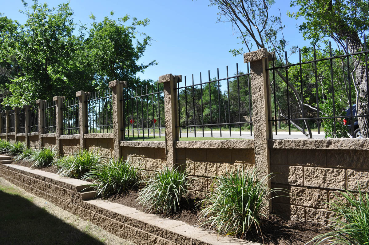 A tan stone retaining wall topped with vertical black metal railings runs alongside a planted bed with green ornamental grasses, with trees and a road visible in the background.