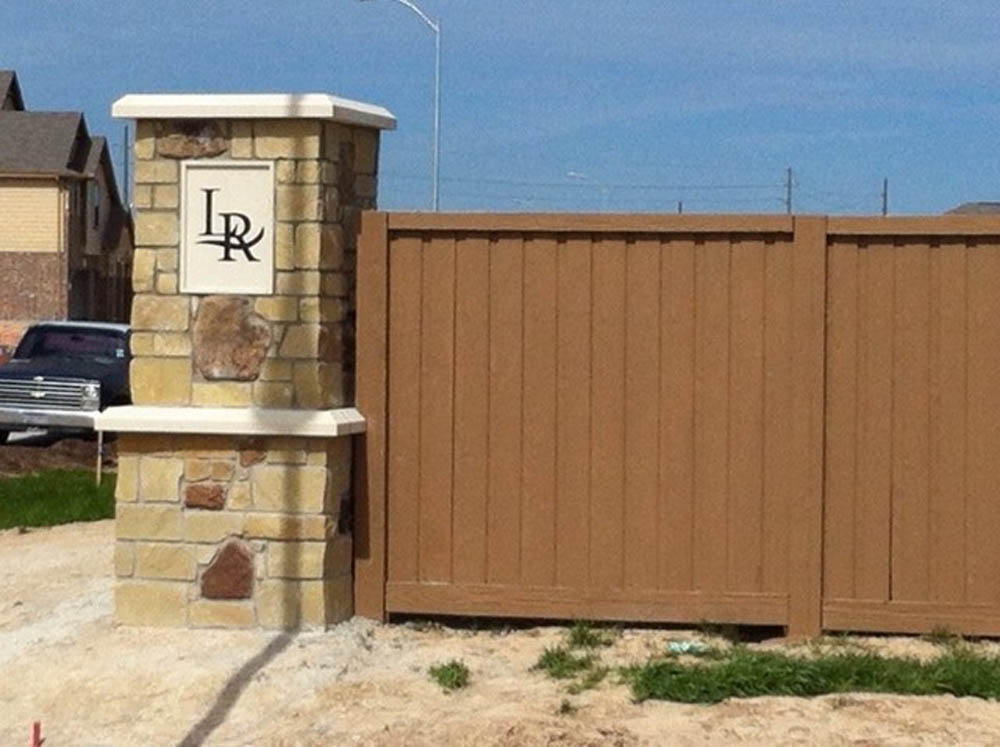 A stone pillar with a plaque showing the letters “LR” stands next to a tall, brown wooden fence near a residential area under a clear sky.
