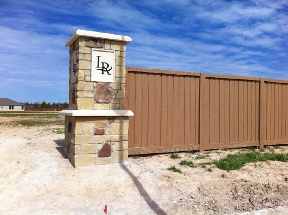 A stone pillar with the black letters LR stands next to a tall brown wooden fence on a dirt lot, under a blue sky with scattered clouds. A house is visible in the distant background.