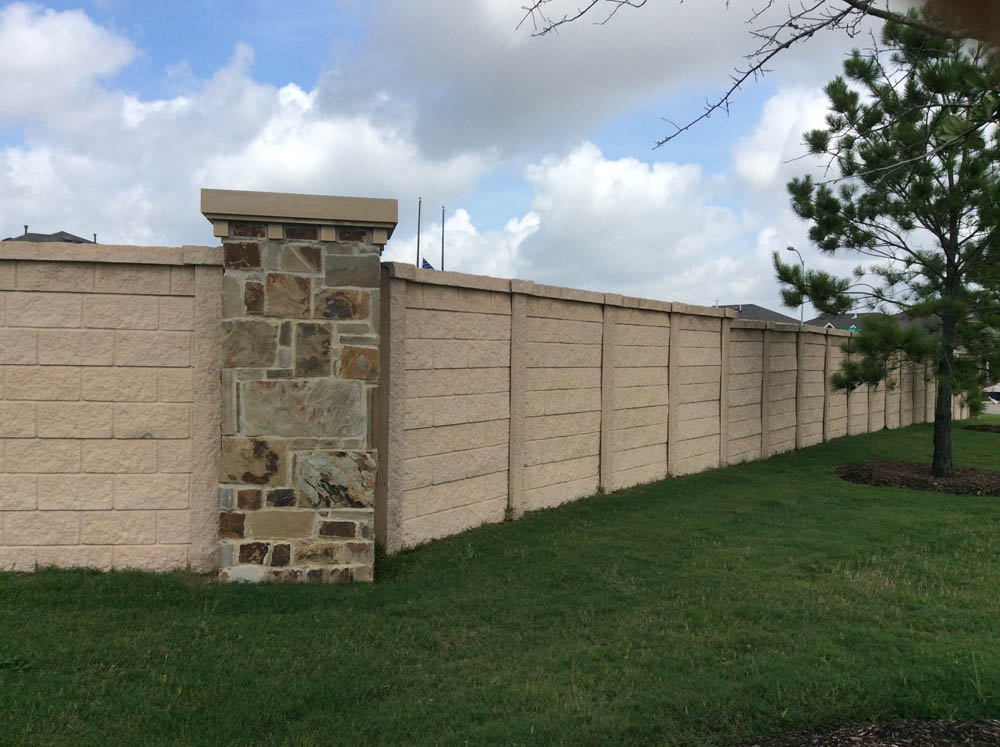 A long beige concrete wall with a decorative stone pillar stands beside a manicured lawn, with a few small trees and a cloudy sky in the background.
