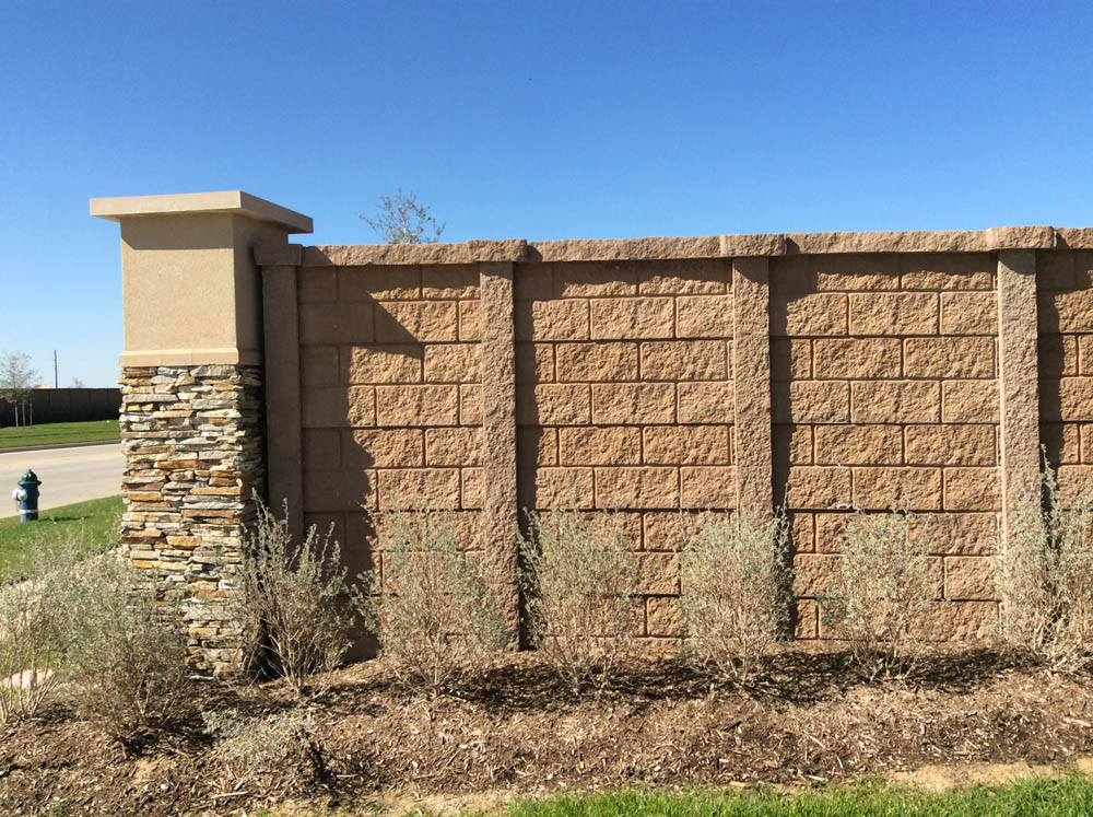 A tan brick and stone wall with square columns stands behind a row of small shrubs, with grass and a blue sky in the background. A fire hydrant is visible on the left side of the image.