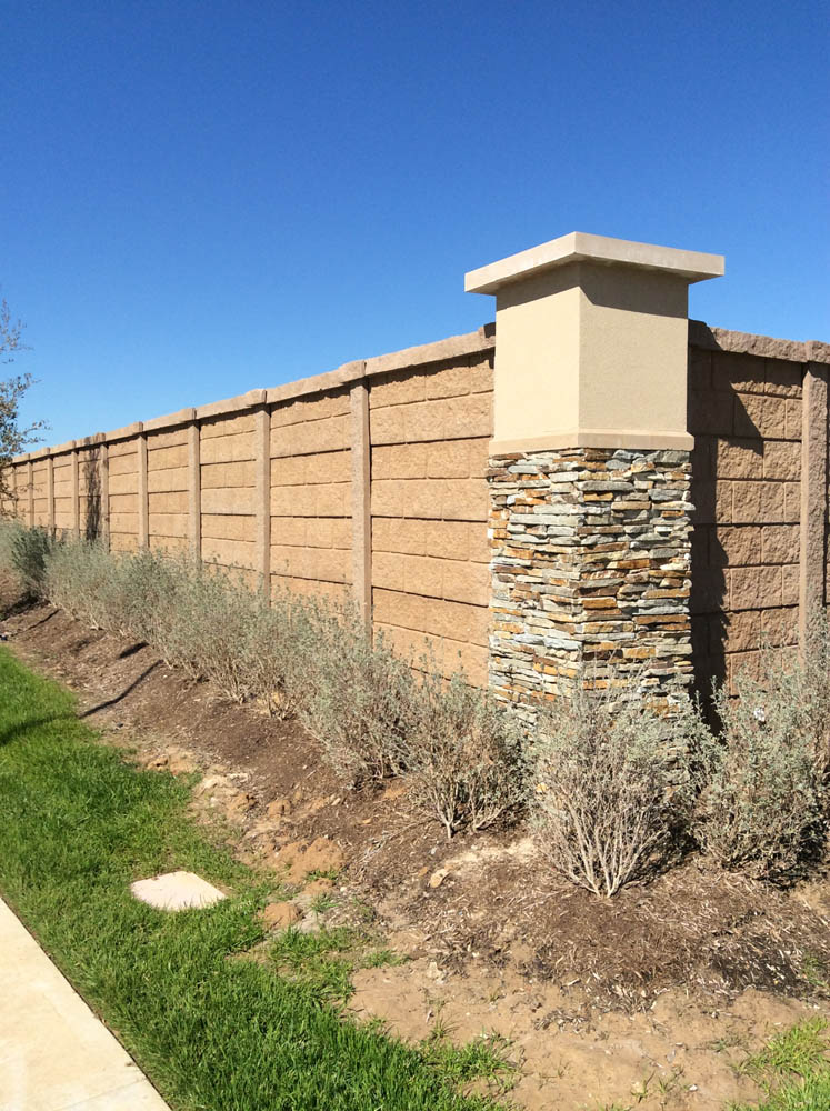 A tall tan brick privacy wall with a decorative stone and stucco pillar runs alongside a landscaped area with small shrubs and green grass under a clear blue sky.