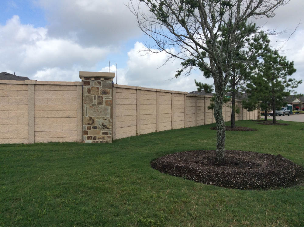 A tall beige stone and concrete privacy wall runs along a grassy area with two mulched trees. Houses and a partly cloudy sky are visible in the background.