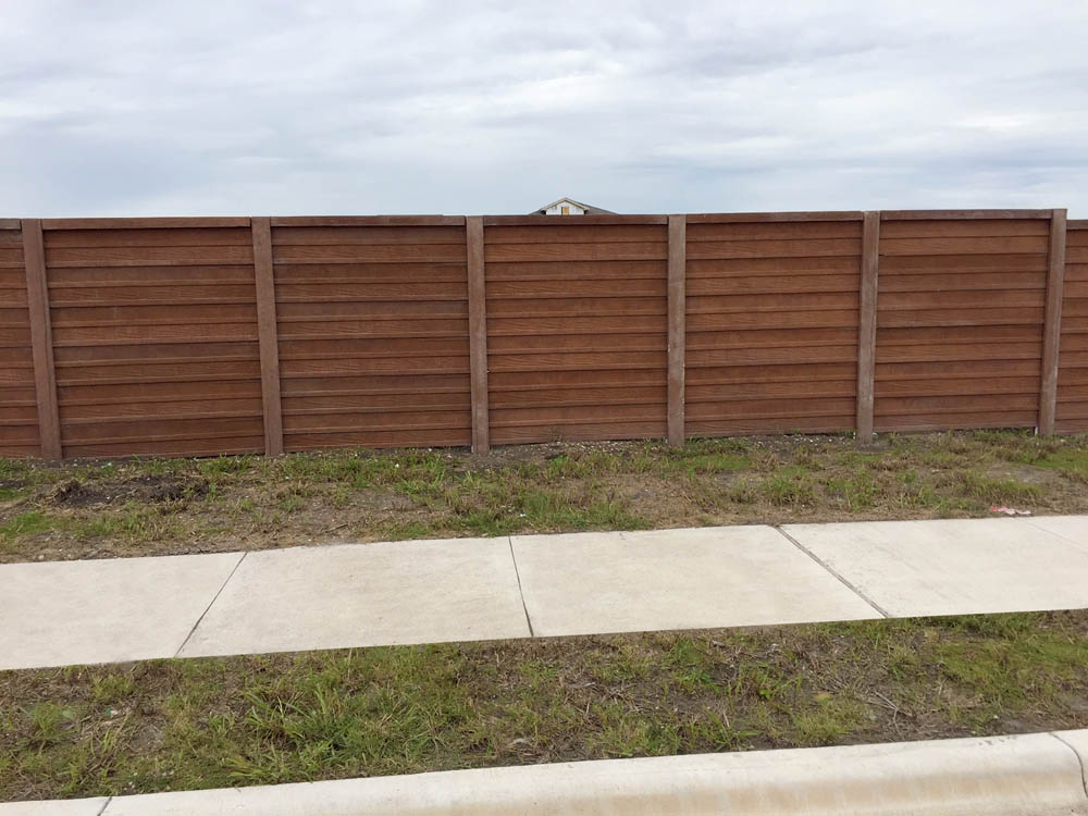 A brown wooden fence stands behind a patch of grass and a concrete sidewalk, with a cloudy sky in the background. Only part of a house roof is visible over the fence.