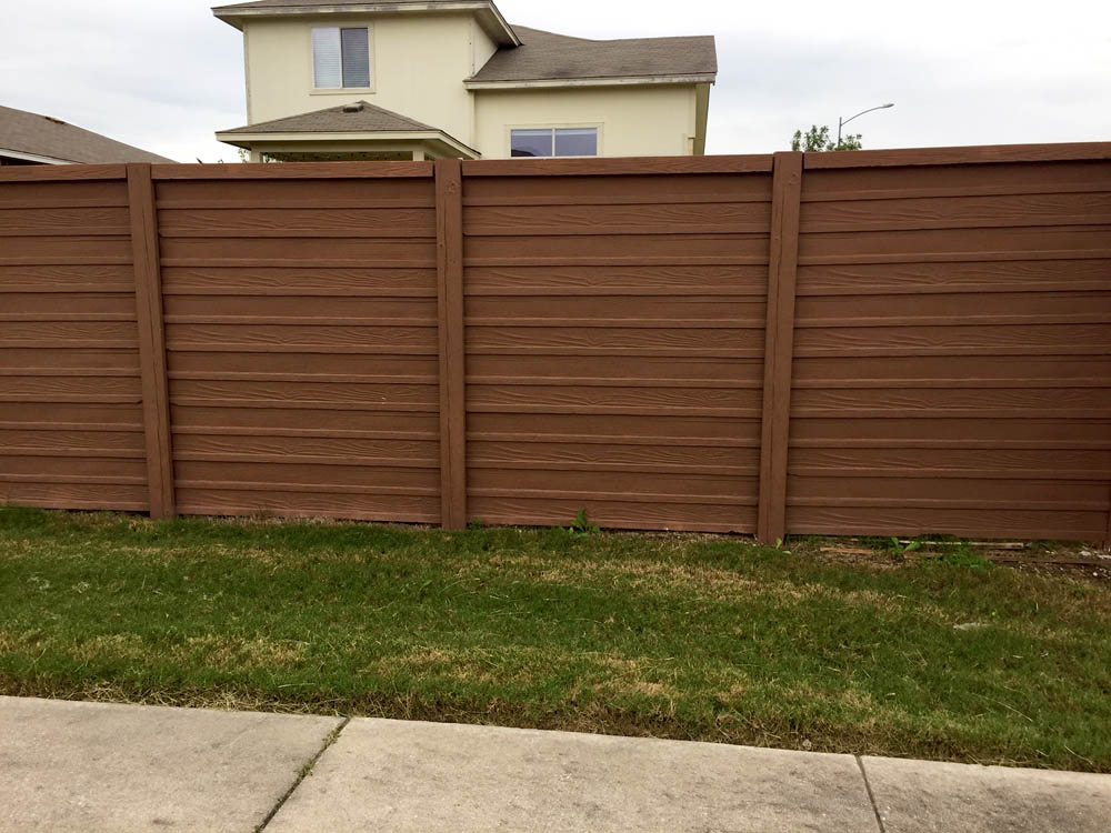 A tall brown wooden privacy fence stands on a grassy yard, with part of a beige two-story house visible behind it. A concrete sidewalk runs along the front of the fence.