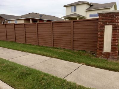 A wooden privacy fence with horizontal panels and brick posts stands alongside a sidewalk and green grass in front of suburban houses.