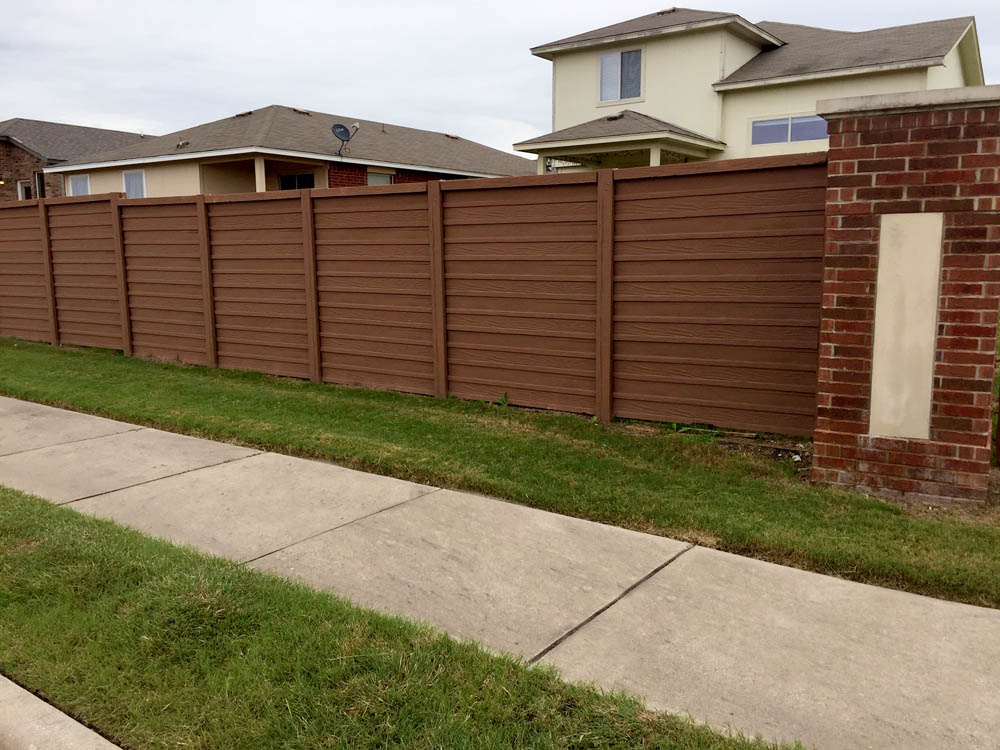 A wooden privacy fence with horizontal panels and brick posts stands alongside a sidewalk and green grass in front of suburban houses.