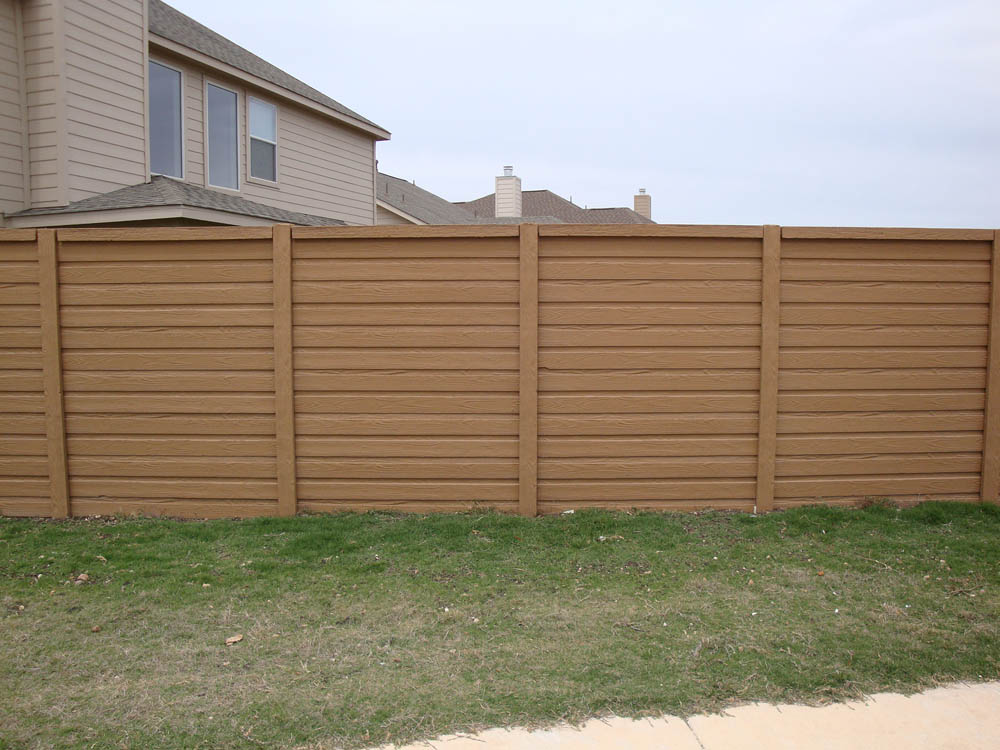 A tall, brown wooden privacy fence stands in front of a suburban house with beige siding. The fence is set on a grassy lawn next to a sidewalk under an overcast sky.