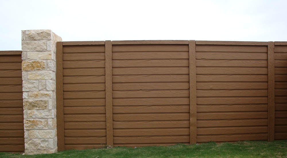 A tall brown horizontal wooden fence with vertical support posts and a stone column on the left side, standing on a patch of green grass under a cloudy sky.