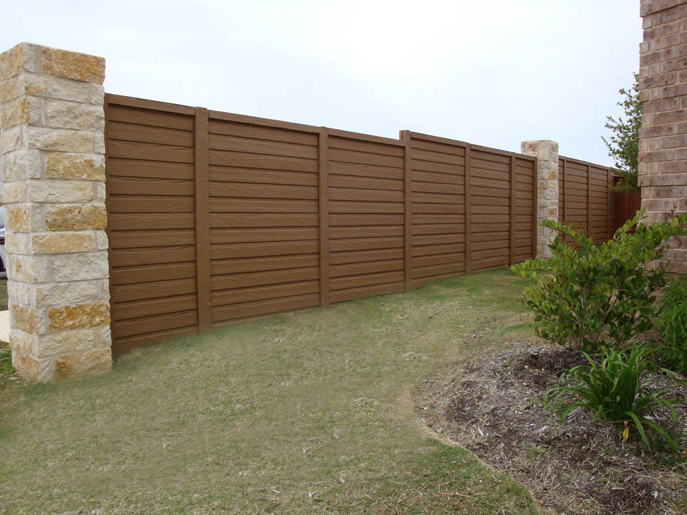 A tall wooden privacy fence with horizontal panels and stone pillars stands on a grassy lawn, with some landscaping and green plants visible in the foreground.