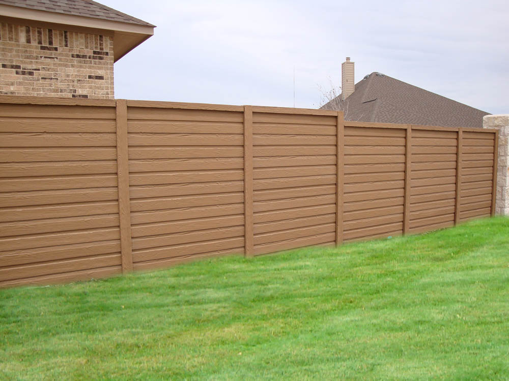 A tall, brown horizontal wooden fence runs along the edge of a neatly mowed green lawn, separating two brick houses under a cloudy sky.