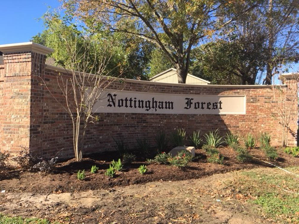 A brick wall sign with the words Nottingham Forest in black letter font, surrounded by small plants and trees, stands under a clear blue sky.