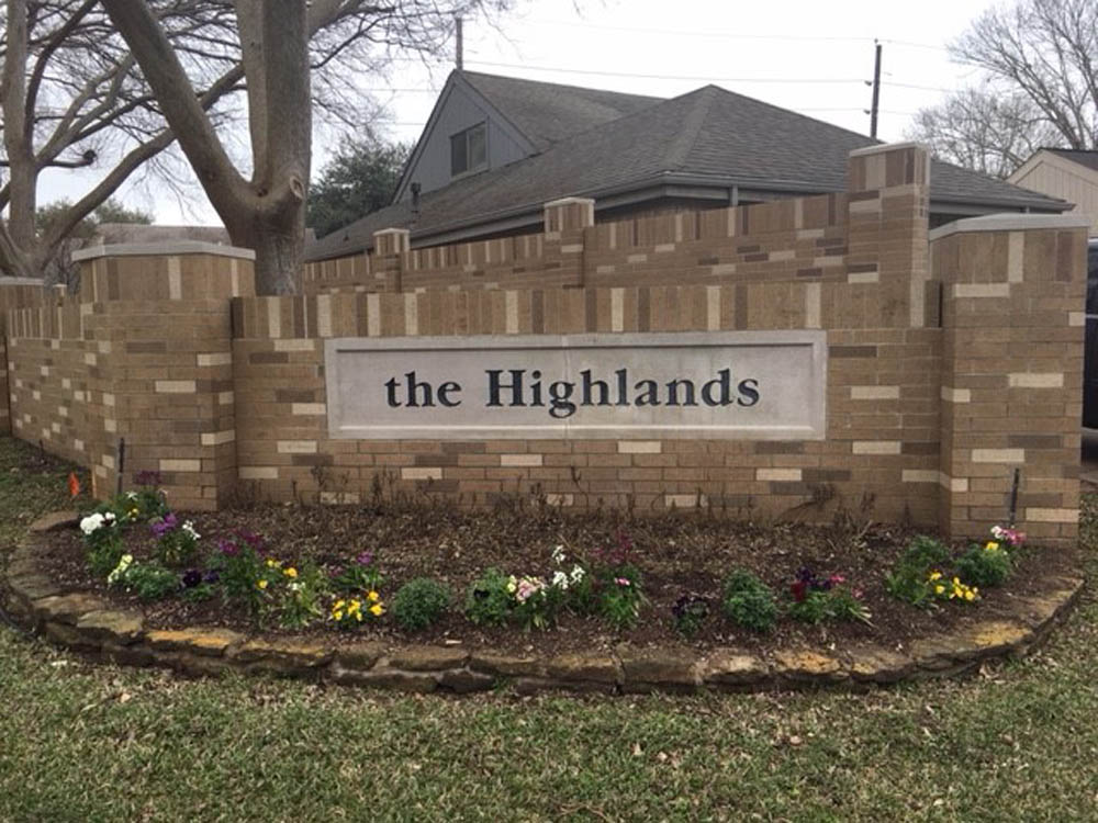 A brick entrance sign reading the Highlands is set in front of a brick wall with a flower bed containing colorful flowers and green plants below it. Trees and a house are visible in the background.