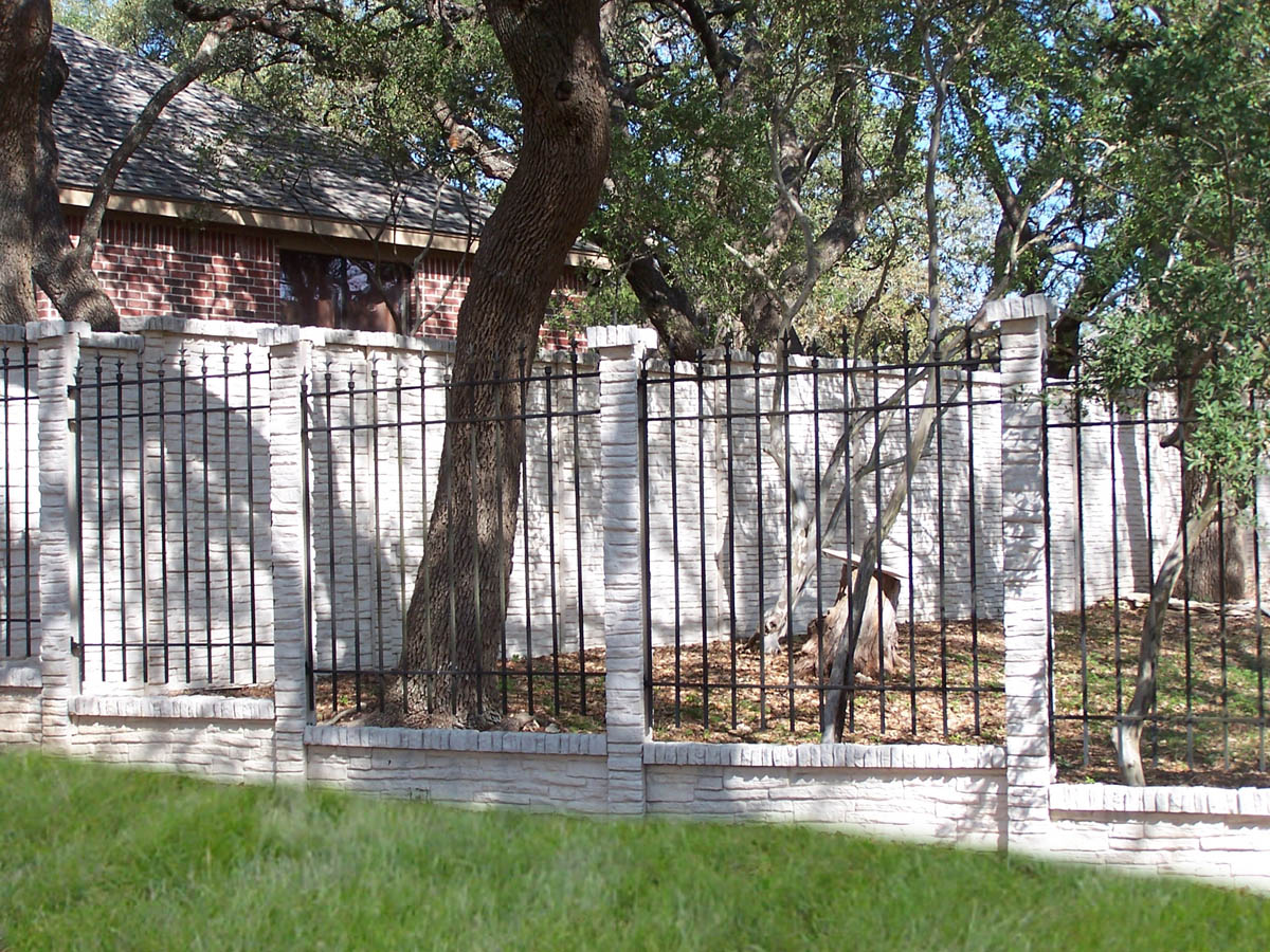 A donkey stands behind a black metal fence with white brick posts and a brick wall, surrounded by trees and a house with red brick and a shingled roof in the background. Grass is visible in the foreground.