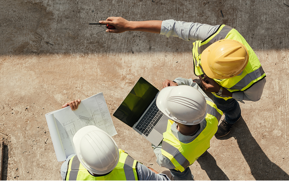 Three construction workers in safety vests and helmets stand outdoors, reviewing building plans and a laptop. One worker points while the others look at the documents, suggesting a discussion about the project.