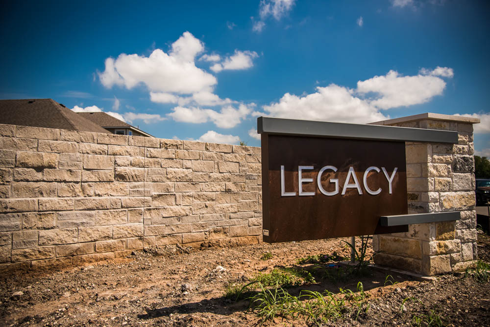 A large stone wall with a metal sign reading LEGACY stands under a bright blue sky with scattered clouds. Some grass and dirt are visible in front of the sign.