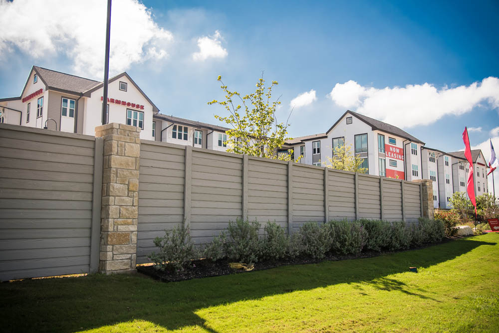 A tall wooden fence with stone pillars runs along a landscaped grassy area, with bushes planted in front. Modern apartment buildings with red and white exteriors are visible in the background under a blue sky with clouds.