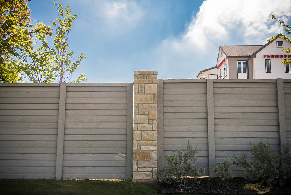 A tall wooden fence with a stone pillar stands in front of a garden, under a blue sky with clouds. Part of a house with a sign reading FARMHOUSE is visible in the background.