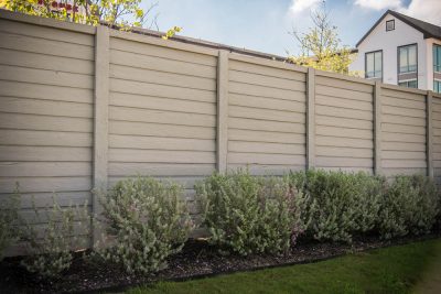 A tall beige privacy fence with horizontal panels stands behind a row of green, bushy shrubs, with grass and a modern house visible in the background.