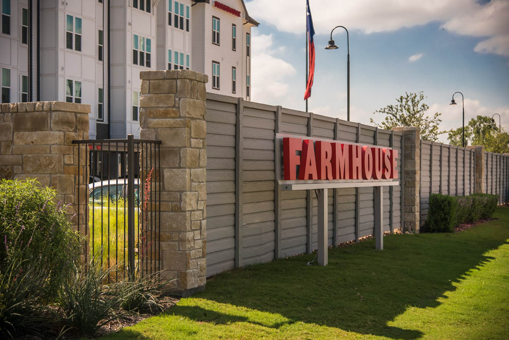 A large sign reading FARMHOUSE in red letters stands in front of a gray wooden fence and stone pillars, with a grassy lawn and modern apartment buildings in the background.