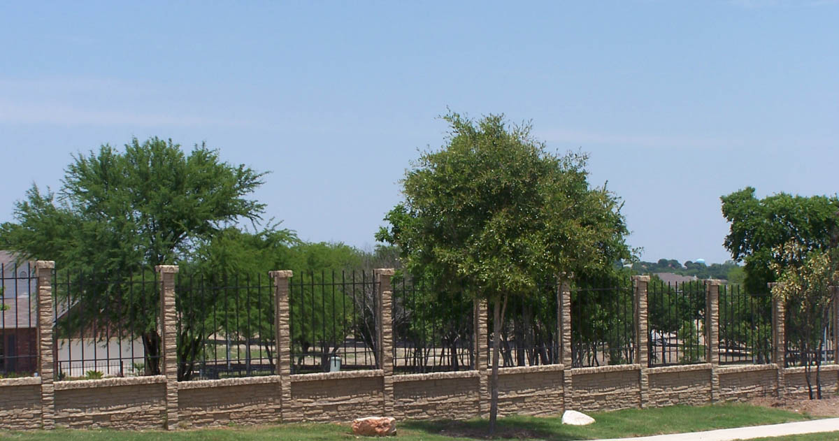 A stone and metal fence stands in front of a row of green trees on a sunny day, with a clear blue sky and houses visible in the background.