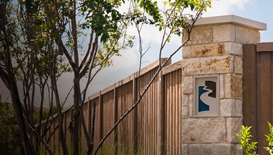 A wooden fence lined with stone pillars, one pillar featuring a small decorative plaque with a blue and brown abstract design, and trees and greenery in the foreground.