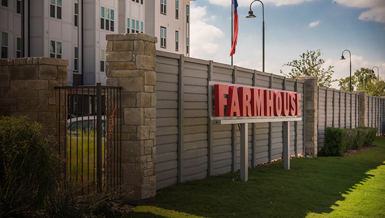 A large red sign reading FARMHOUSE is mounted on a wooden fence with stone columns in front of an apartment building. A gate is on the left, and a U.S. flag flies overhead. Grass and landscaping line the fence.