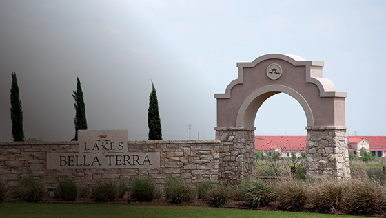 Stone entrance sign for Lakes of Bella Terra with an arched gateway, trimmed bushes, and tall grasses in front, and a building with a red roof visible in the background.