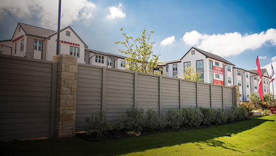 A tall, gray privacy fence with stone pillars stands in front of modern apartment buildings under a partly cloudy sky, with shrubs and well-kept grass in the foreground.