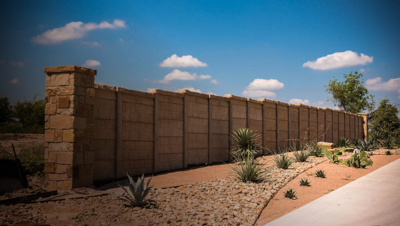 A tall stone and concrete wall runs alongside a landscaped area with desert plants, rocks, and a paved path under a bright blue sky with scattered clouds.