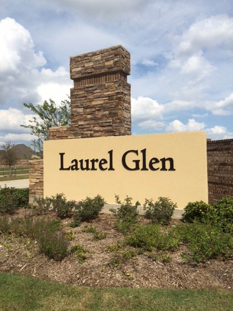 A stone and brick entrance sign reads Laurel Glen in large black letters, set against a light-colored background with landscaping and a cloudy sky in the background.