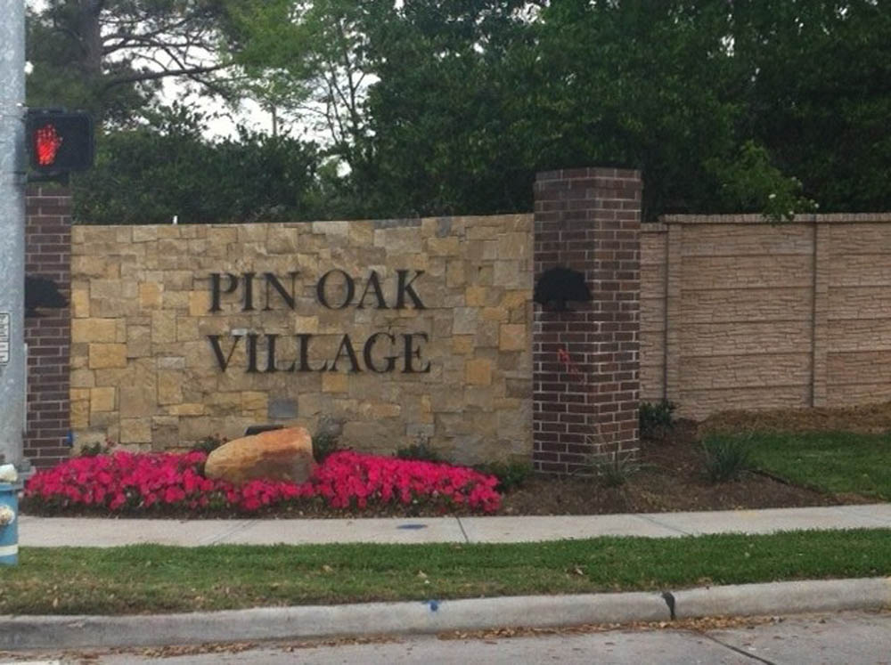 A stone and brick entrance sign reads PIN OAK VILLAGE, with pink flowers, a large rock, and greenery in front; trees and a sidewalk are also visible.