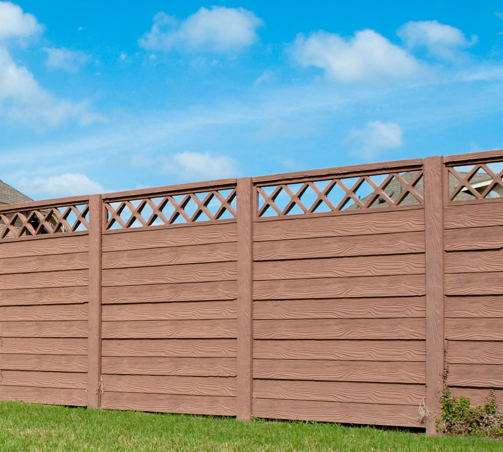 A tall wooden privacy fence with a lattice top section stands on green grass under a bright blue sky with scattered white clouds.