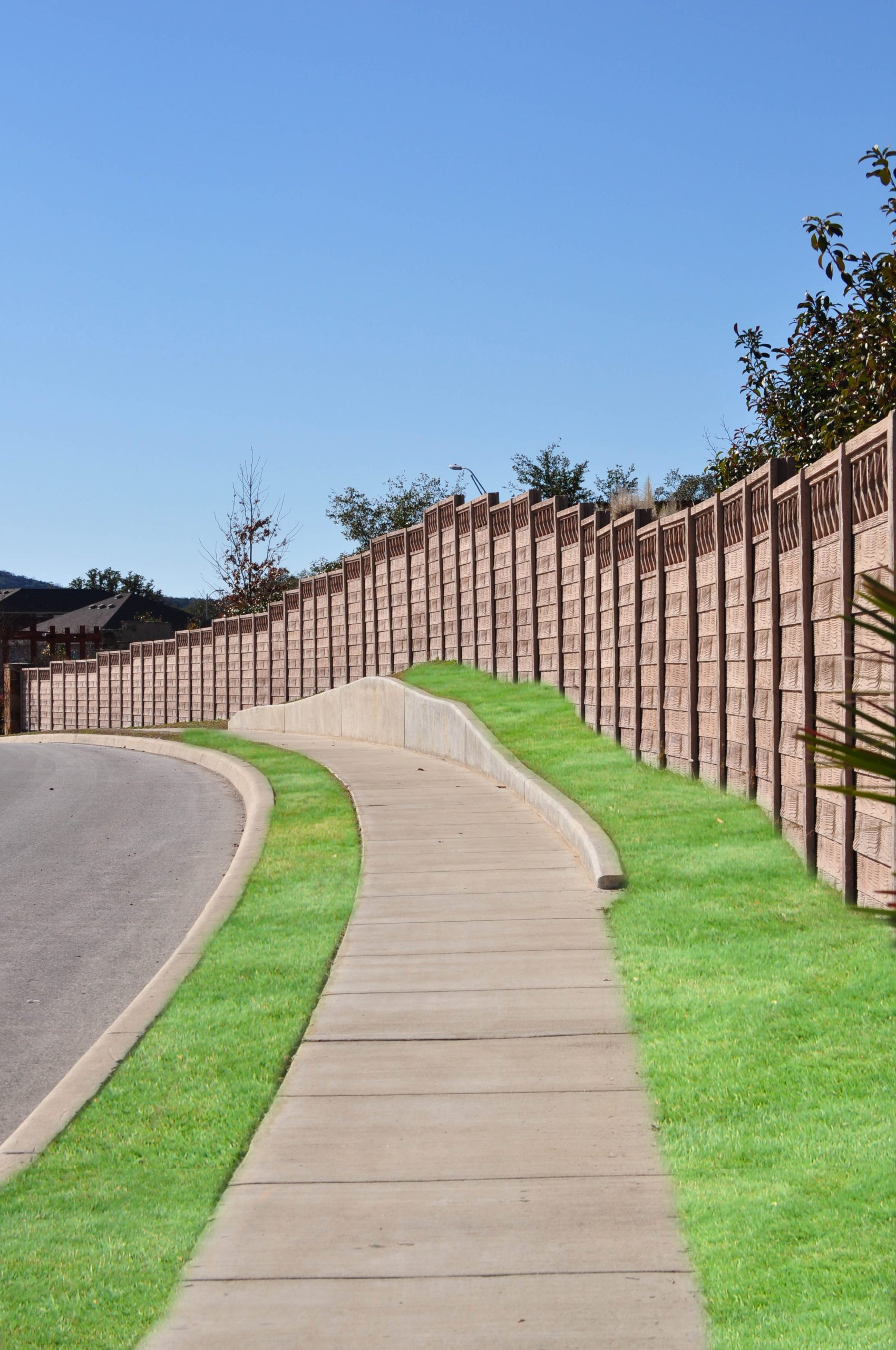 A concrete sidewalk curves beside a paved road, bordered by bright green grass and a tall wooden fence under a clear blue sky. Trees and distant hills are visible in the background.