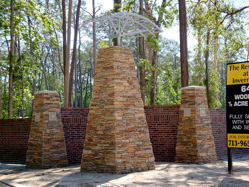 Three stone pillars with decorative metalwork stand in front of a brick wall, surrounded by tall trees. A black and yellow real estate sign is partially visible on the right.