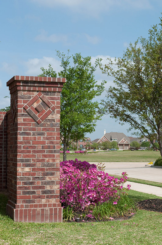 A brick column with a diamond pattern stands near blooming pink flowers and green trees; in the background, houses line a suburban street under a blue sky with scattered clouds.