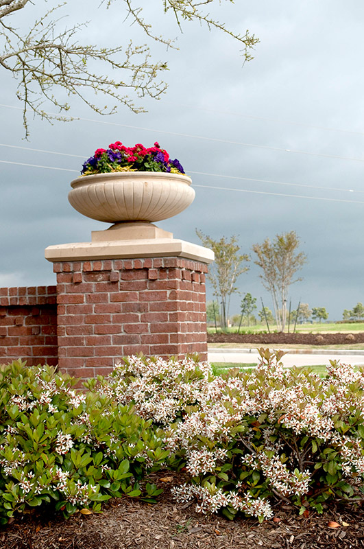 A large stone planter filled with colorful flowers sits atop a red brick column, surrounded by blooming shrubs and greenery, with cloudy skies and open landscape in the background.