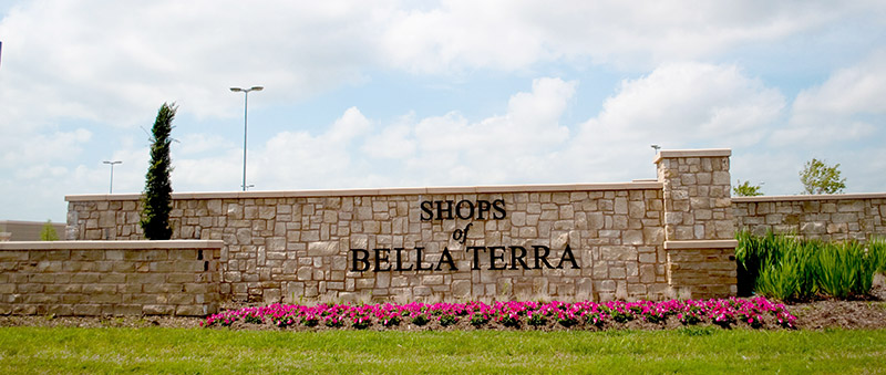 A stone sign reads Shops of Bella Terra in black letters, surrounded by a flower bed with pink flowers and green grass, under a partly cloudy sky.