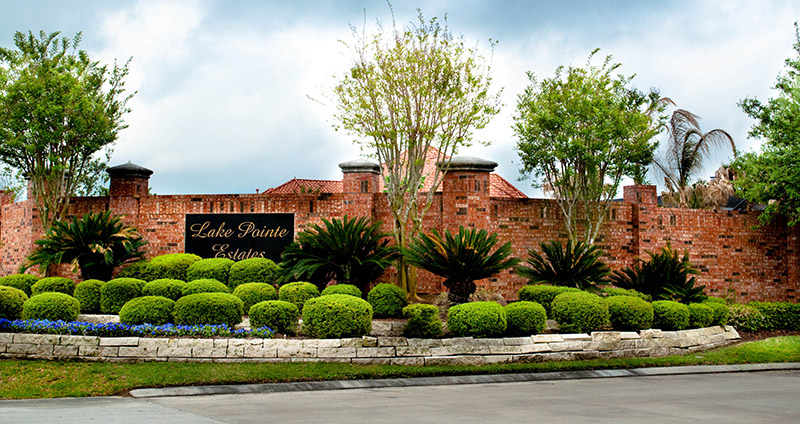 A red brick entrance sign for Lake Pointe Estates is surrounded by neatly trimmed bushes, flowers, palm plants, and trees, with a stone border along the sidewalk and a cloudy sky above.