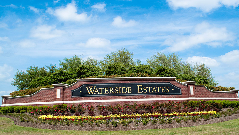 A brick entrance sign with gold letters reads Waterside Estates. The sign is surrounded by neatly trimmed bushes, colorful flowers, and a backdrop of green trees under a blue sky with scattered clouds.
