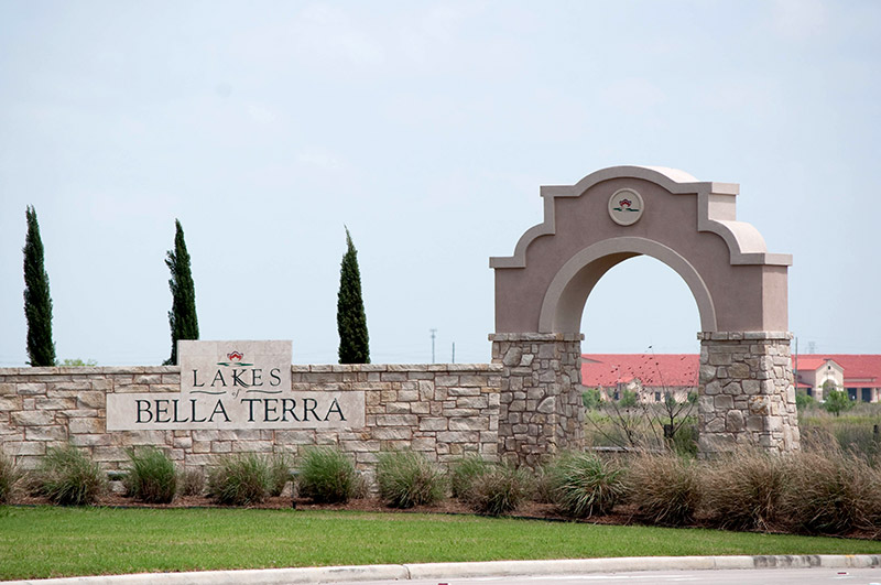 Entrance sign for Lakes of Bella Terra featuring a stone wall with lettering and an arched architectural structure, landscaped with grass and tall bushes, under a partly cloudy sky.
