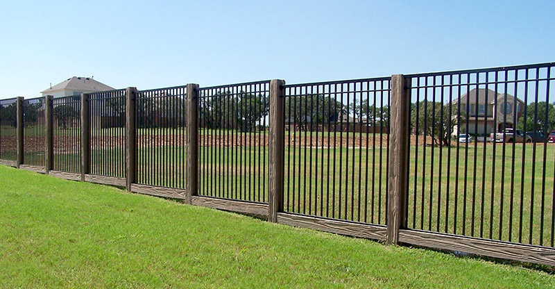 A black metal fence with wooden posts runs along a grassy field, with houses and trees visible in the background under a clear blue sky.