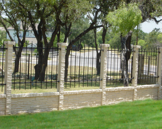 A stone and wrought iron fence runs along a grassy area with several trees behind it, and a paved road is visible in the background.