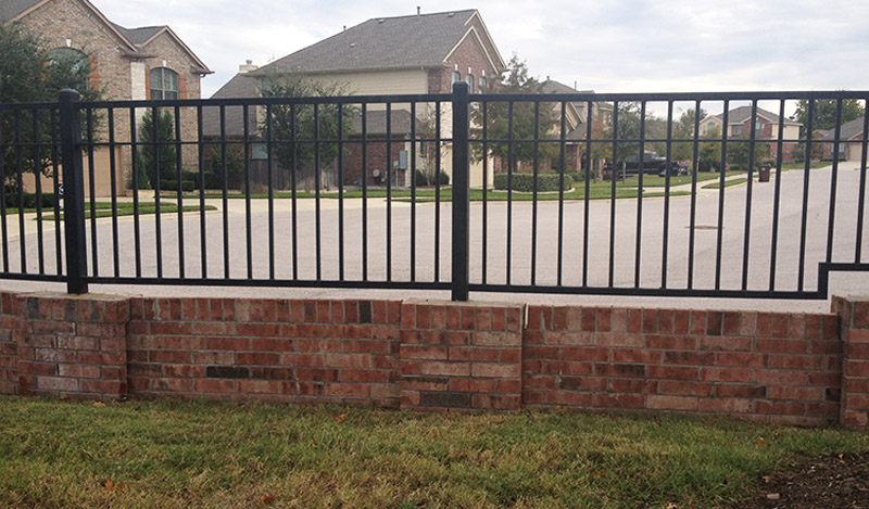 A black metal fence with vertical bars is mounted on a short red brick wall, separating a grassy yard from a suburban street with houses in the background.