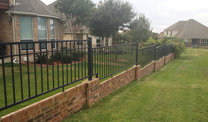A black metal fence with brick base separates two grassy yards between suburban houses with sloped roofs and large windows. Trees and shrubs line the fence.