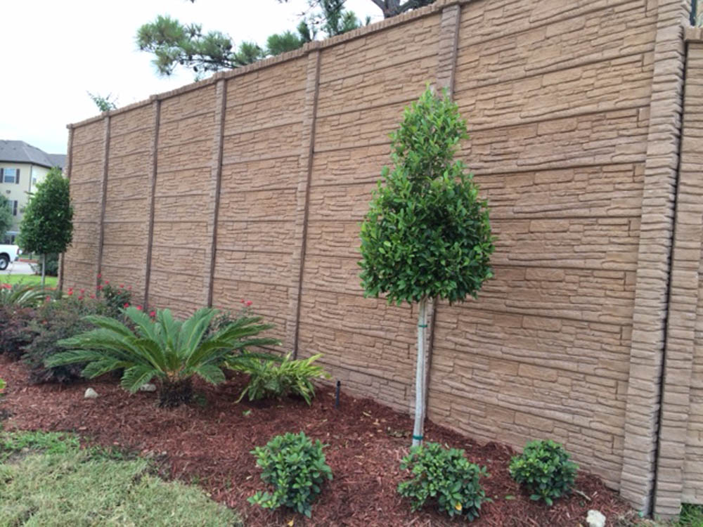 A landscaped garden with a small tree, shrubs, and mulch in front of a tall, textured brown stone wall. Green plants are neatly arranged along the base of the wall.
