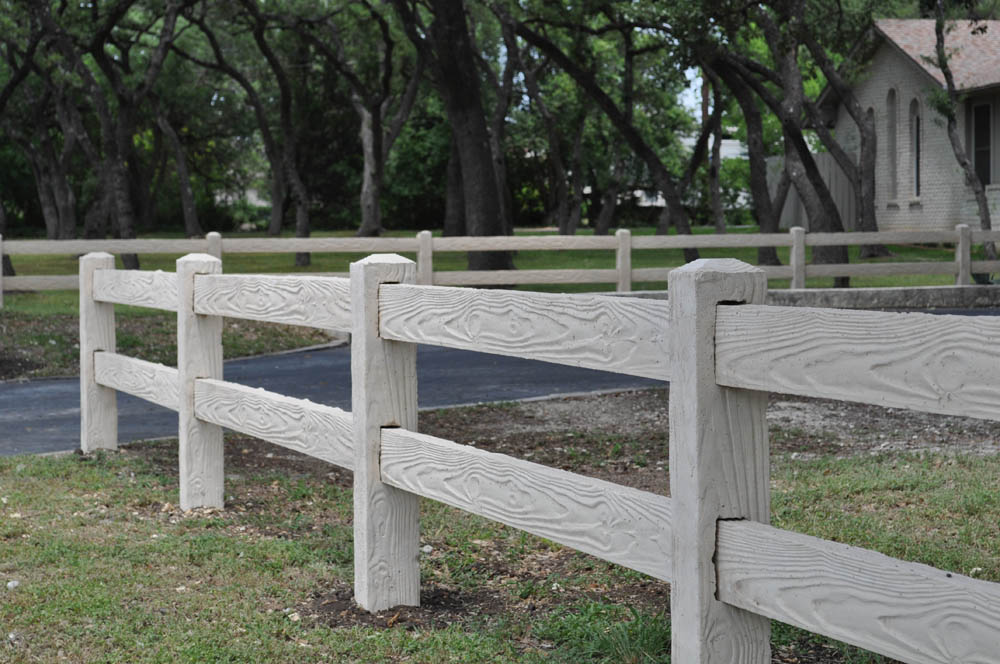 A close-up of a light-colored wooden fence with two horizontal rails and vertical posts, set next to a grassy area with trees and a house in the background.